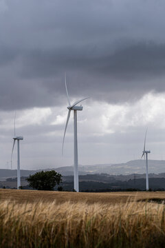 Three wind turbines dominate a field under a stormy sky, highlighting the blend of modern renewable energy technology amid rural landscapes and the power of natural elements.