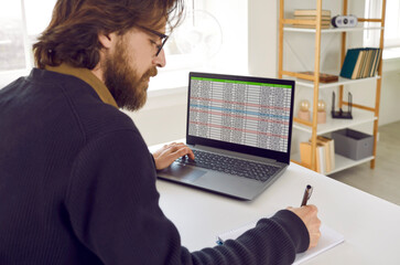 Male entrepreneur or business accountant working on laptop computer. Bearded man sitting at office desk with notebook PC, working with database, using data sheets, writing down information in notepad