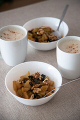 A cozy breakfast setting with two white cups of frothy cappuccino and two bowls of baked apple, nuts and pumpkin seeds on a beige tablecloth, perfect for a relaxed and healthy morning.