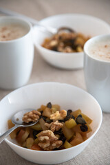A cozy breakfast setting with two white cups of frothy cappuccino and two bowls of baked apple, nuts and pumpkin seeds on a beige tablecloth, perfect for a relaxed and healthy morning.