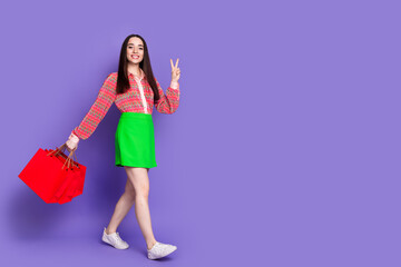 Full size photo of pretty young girl walking shopping bags v-sign wear trendy pink outfit isolated on purple color background