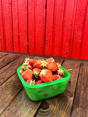 Strawberries are in a green container, on a wooden bench, against a red wall