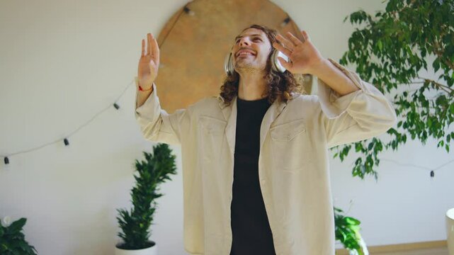 A young man is enjoying music with wireless headphones, smiling, and dancing in a cozy home environment. He seems happy and carefree, showing joyful expression while listening to music in his