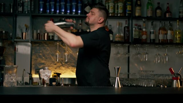 Young male bartender apron preparing alcohol drink shaking it in cocktail shaker standing in front of bar counter