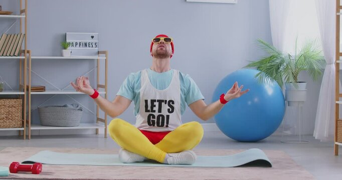 Humorous yoga. Cheerful eccentric man meditating funny and breathing deeply while sitting on yoga mat in room. Man is wearing funny colored sportswear, red headband and sunglasses. 4k footage.