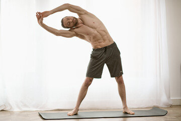 Handsome man standing on yoga mat, practicing yoga in front of window.