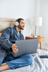 Man on bed, in headphones, laptop in use.