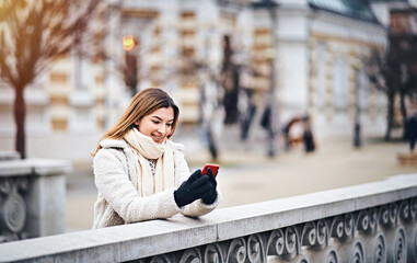 Millennial woman in jacket relying on a stone railing while typing an online message on her mobile phone with gloves on here hand - Outdoor smartphone usage concept in cold winter weather