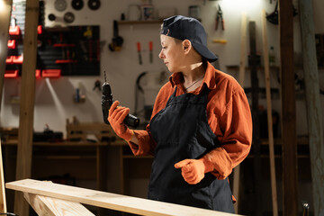 Middle-aged woman carpenter with electric drill working in her woodworking workshop