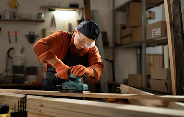 Female carpenter in safety glasses sanding wooden board using belt sander, working in her workshop.