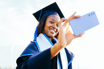 African woman in a graduation gown is taking a picture of herself with a cell phone.