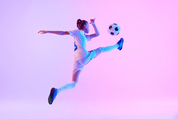 Dynamic image of focused teen girl, soccer player in motion, practicing, kicking ball in jump against pink studio background in neon. Concept of sport, active and healthy lifestyle, childhood, school