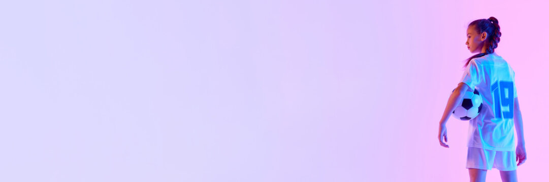 Teen girl, soccer player in white uniform posing with ball against pink studio background in neon light. Concept of sport, active and healthy lifestyle, childhood, school, hobby. Banner for ad - Powered by Adobe