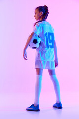 Full-length image of teen girl in white uniform, football player posing with ball against pink studio background in neon light. Concept of sport, active and healthy lifestyle, childhood, school, hobby