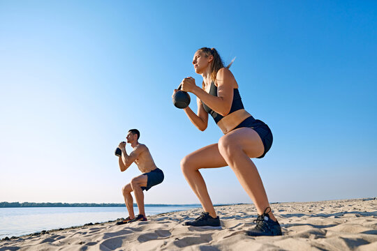Sportive young man and woman with sportive body, in black sportswear doing squats with kettlebell, intensive outdoor training on beach by the river. Concept of sport, healthy lifestyle, body care