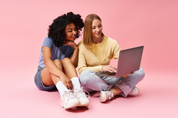 Two diverse women in casual attire sitting on floor, engrossed in laptop.