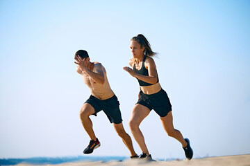 Dynamic and hard training outdoor session. Sportive young man and woman training on sandy beach, running under clear sky. Concept of sport, active and healthy lifestyle, body care, fitness