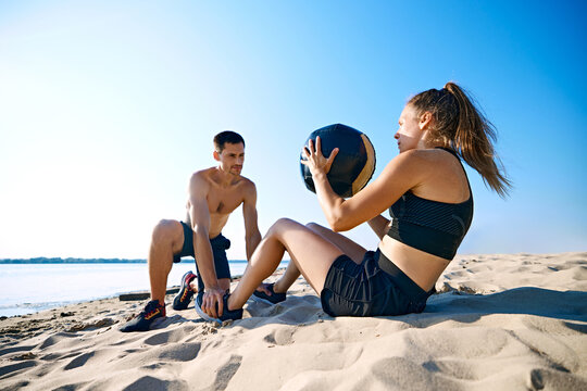 Man helping woman with hard press exercises with medicine ball on sandy beach under clear blue sky. Intensive workout. Concept of sport, active and healthy lifestyle, body care, fitness - Powered by Adobe