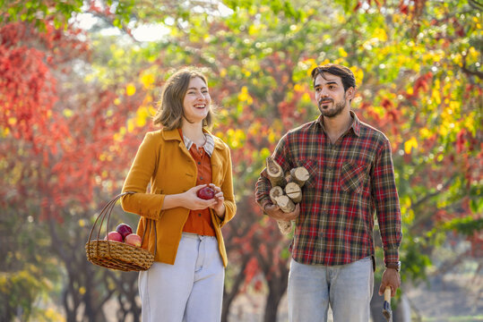 young couple farm workers,a female holding a basket of apples,a man carry firewood and axe while walking home ,concept of seasonal harvesting in autumn - Powered by Adobe