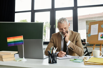 A middle-aged man with a beard sits at a desk with a laptop and a rainbow flag, thoughtfully writing in a notebook.