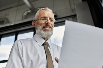 A mature man with a beard, wearing a white shirt and tie, teaches from a bright office.