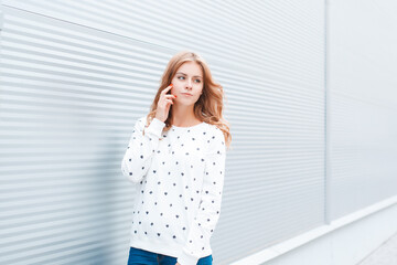 Beautiful fashion young happy woman with natural red hair in a stylish white sweater near a yellow modern building in the city