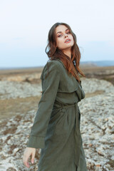 Mysterious woman in trench coat standing on rocky hillside with hands in pockets on overcast day