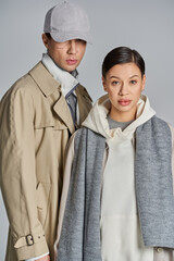 A young stylish couple wearing trench coats standing together in a studio against a grey background.