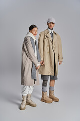 Young stylish couple in trench coats standing side by side in a studio, exuding urban elegance against a grey background.