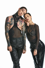 A young stylish and tattooed man and woman strike a pose together in a studio against a grey background.