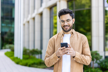 Smiling young man wearing glasses using smartphone outside modern office building. Casually dressed man engaging with social media or business apps phone, bright environment representing technology