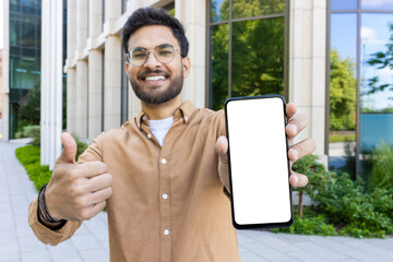 Smiling man in brown shirt and glasses holding smartphone with blank screen and showing thumbs up gesture outside modern building with greenery and glass windows
