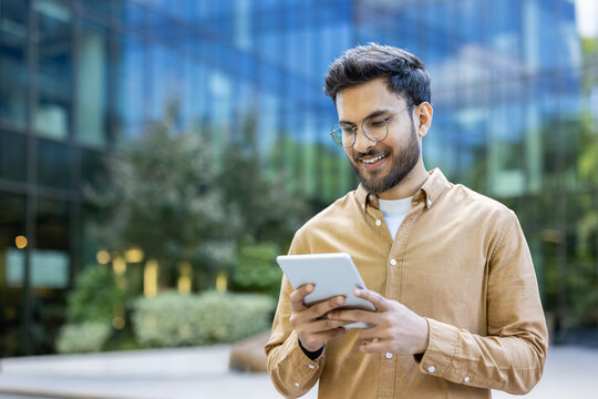 Young man smiling while using tablet outside modern office building. Casual attire, urban setting, and contemporary design. Represents technology, business, and communication. Happy expression - Powered by Adobe