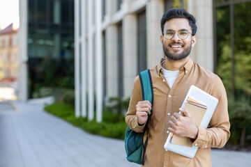 Confident student standing outside modern university building. Young man wearing glasses, carrying backpack, holding books and laptop. Concept of education, college life, academic success, study