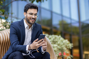 Young businessman sitting on bench listening to music with earphones using smartphone outdoors. Wearing suit and glasses, smiling and relaxed, office building in background.