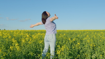 Fototapeta premium Woman Standing In Yellow Rapeseed Field Raising Her Arms Expressing Freedom. Beautiful Healthy Caucasian White Woman Standing In Blooming Rapeseed Field And Looking At Horizon.