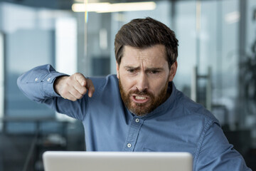 An angry young man in a blue shirt and with a beard is sitting in the office at a desk in front of...