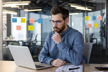Young serious male businessman wearing glasses sitting in modern office at workplace, looking at...