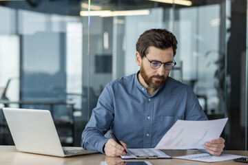 Concentrated and serious young male accountant sitting in the office behind a desk and working with documents, checking and analyzing information and data