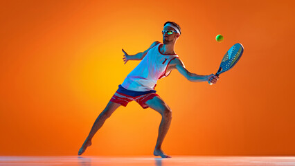Young man, tennis player in uniform and reflective sunglasses playing beach tennis against orange...