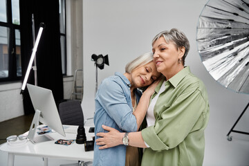 A mature lesbian couple, one a photographer and the other a model, share a tender embrace in a studio setting.