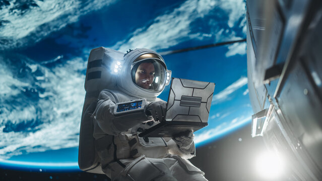 Male Astronaut Using a Laptop to Wirelessly Connect to a Satellite. Spaceman Working on a Device in Open Space Outside Earth Atmosphere. Professional Engineer Undertaking Service and Maintenance Job
