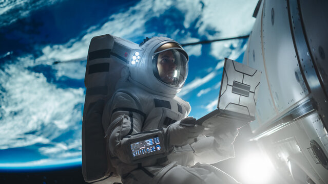Man in a Space Suit Casually Working on a Laptop Computer in Outer Space While Servicing a Satellite. Astronaut Writing to Mission Control, Family or Work Colleagues, Reading Maintenance Protocol
