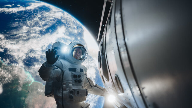 Portrait of Young Astronaut Posing in a Space Suit During a Spacewalk Outside a Spaceship, Satellite or a Station. Spaceman Looking at Camera, Smiling and Waving Hello Next to an Entrance Hatch
