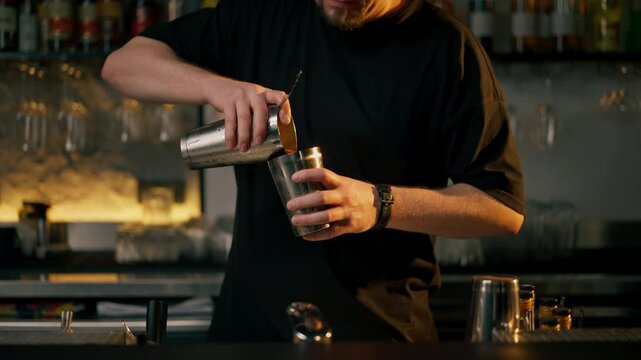 Young male bartender in apron preparing alcohol drink pouring alcohol into cocktail shaker