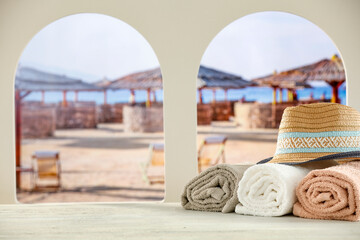 Towels and straw summer hat on wooden table top with copy space for products. Arched windows and blurred summer tropical beach background