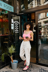A fashionable woman in pink and white pants smiles as she steps out of a European cafe.