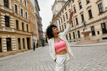 A woman in a white blazer and pink crop top poses on a European street.