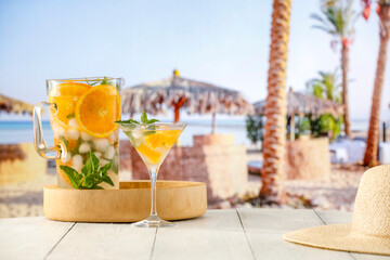Large pitcher of lemaonade mocktail drink with orange and ice and mint leaves on wooden table with tropical beach background.