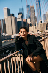 A young woman sits on a railing in New York City, lost in thought amidst the bustling cityscape.